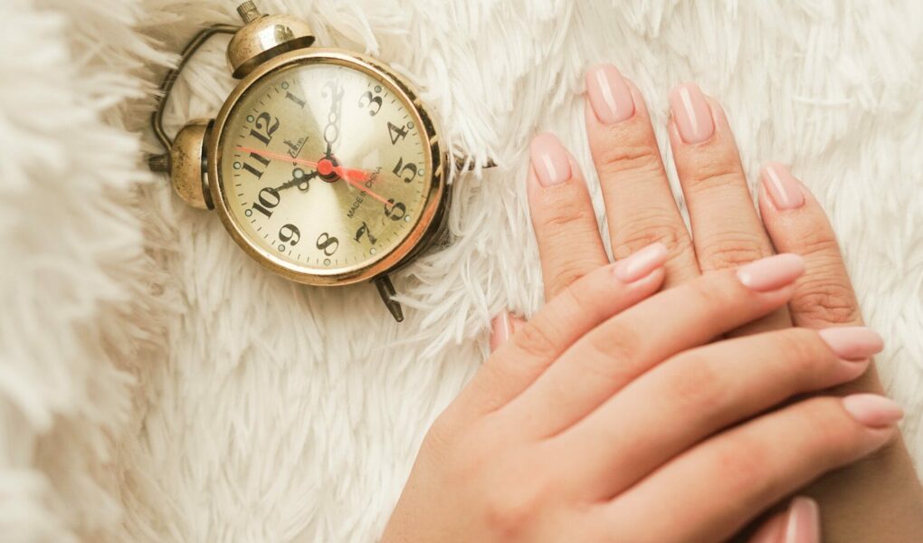 Mains soignées avec une manucure en gel rose nude longue tenue posées à côté d'un réveil rétro