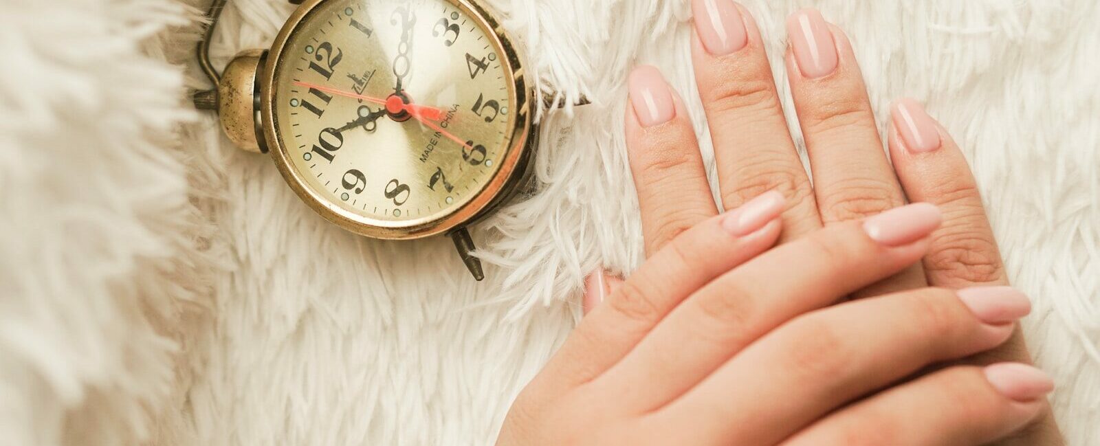 Mains soignées avec une manucure en gel rose nude longue tenue posées à côté d'un réveil rétro