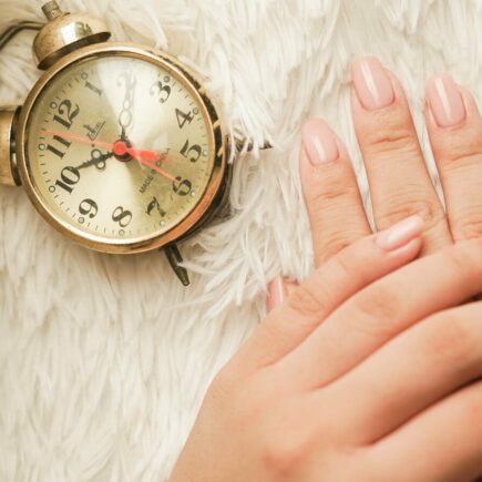 Mains soignées avec une manucure en gel rose nude longue tenue posées à côté d'un réveil rétro