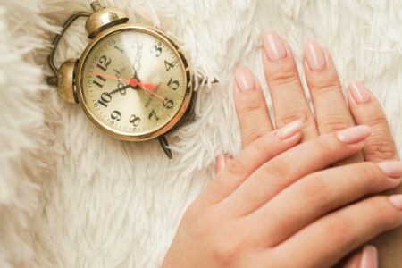 Mains soignées avec une manucure en gel rose nude longue tenue posées à côté d'un réveil rétro